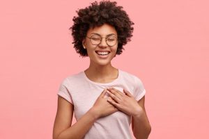 photo of friendly positive female smiles sincerely has afro hair keeps both palms on heart dressed in casual t shirt isolated over pink wall expresses good feelings and attitude to close person